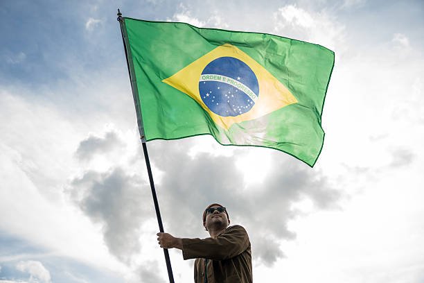 istock Portrait of cheerful young man looking forward while standing proudly with holding Brasil flag over his head against wind and blue sky, Man holding the Brasil flag celebration 2232649063
