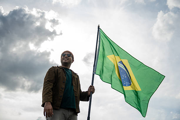 istock Portrait of cheerful young man looking forward while standing proudly with holding Brasil flag over his head against wind and blue sky, Man holding the Brasil flag celebration 2232649069