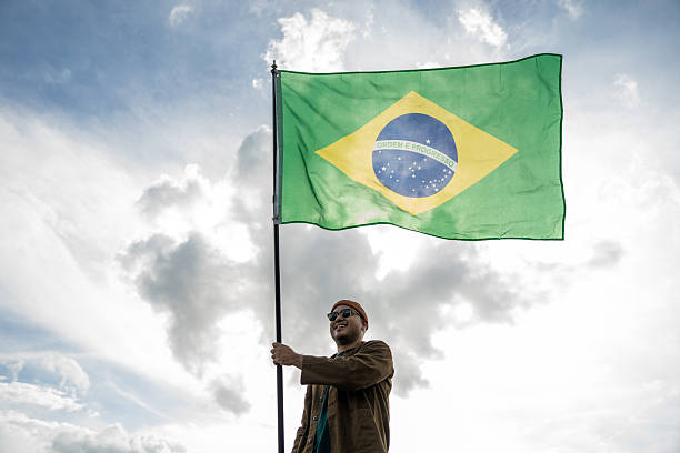 istock Portrait of cheerful young man looking forward while standing proudly with holding Brasil flag over his head against wind and blue sky, Man holding the Brasil flag celebration 2232649101