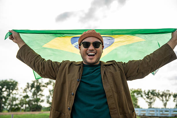 istock Portrait of cheerful young man looking forward while standing proudly with holding Brasil flag over his head against wind and blue sky, Man holding the Brasil flag celebration 2232649145
