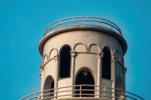 Close up of a vintage lighthouse tower against a clear blue sky, featuring architectural details like arched windows and metal railings. The structure shows signs of age and weathering.