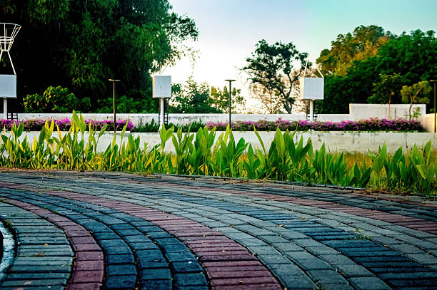 Curved brick pathway in a park with lush green plants and trees. The pathway leads to a white wall with purple flowers and lamp posts. The scene captures nature and architecture.