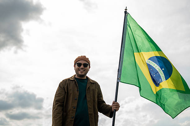 istock Portrait of cheerful young man looking forward while standing proudly with holding Brasil flag over his head against wind and blue sky, Man holding the Brasil flag celebration 2232655170