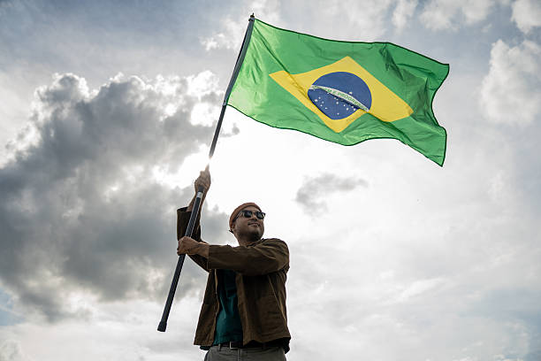 istock Portrait of cheerful young man looking forward while standing proudly with holding Brasil flag over his head against wind and blue sky, Man holding the Brasil flag celebration 2232655177
