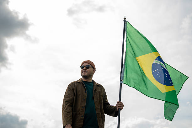 istock Portrait of cheerful young man looking forward while standing proudly with holding Brasil flag over his head against wind and blue sky, Man holding the Brasil flag celebration 2232655184