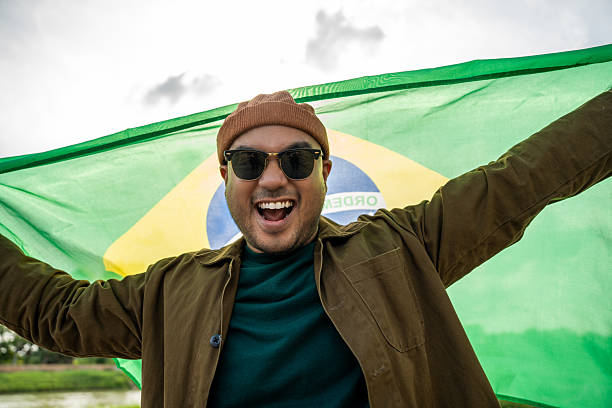 istock Portrait of cheerful young man looking forward while standing proudly with holding Brasil flag over his head against wind and blue sky, Man holding the Brasil flag celebration 2232655206