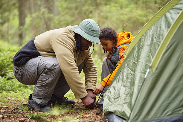 Black man and Black child setting up tent together in forest, crouching and working as team, focusing on securing tent pegs, engaging in outdoor camping activity surrounded by greenery