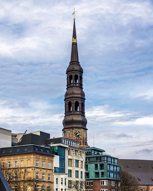 St. Michael's Church tower in Hamburg, Germany, rises above modern and historic buildings under a cloudy sky.