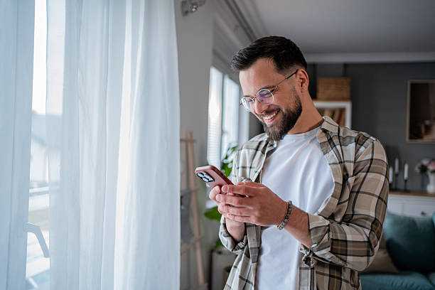istock Smiling young man using smartphone near window at home 2232671688