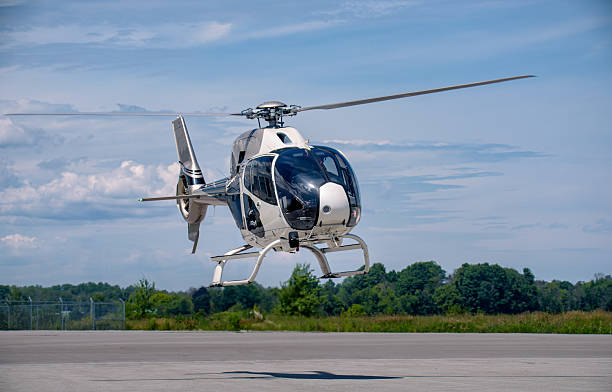 A white Airbus H120 helicopter is captured in a low hover above the runway. The aircraft’s landing skids are visible in detail, and its main rotor shows subtle motion blur. In the distance, a line of green trees frames the horizon, while the sky is filled with layered alto-stratus and cirrus clouds, creating a textured and atmospheric backdrop.