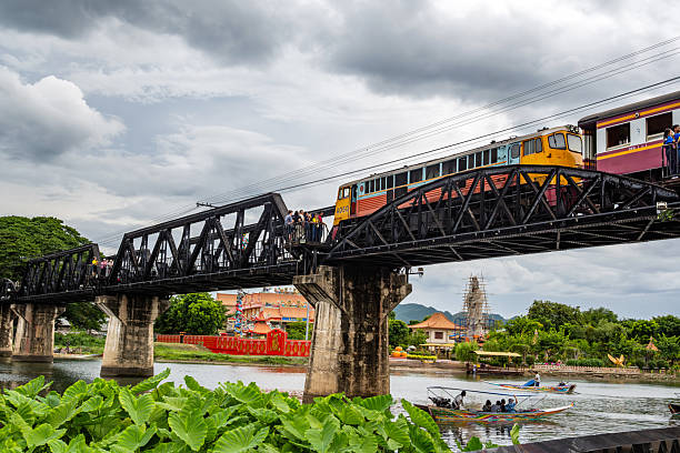 Scenic view of a train traversing the historic Bridge on the River Kwai, surrounded by lush greenery and vibrant riverside activity. The image captures an iconic cultural and historical landmark in Kanchanaburi, Thailand.