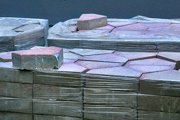 Stacks of rectangular concrete paving blocks arranged neatly on wooden pallets in an outdoor storage yard. The gray tones and geometric alignment reflect construction efficiency and industrial organization, with visible textures on the block surfaces.