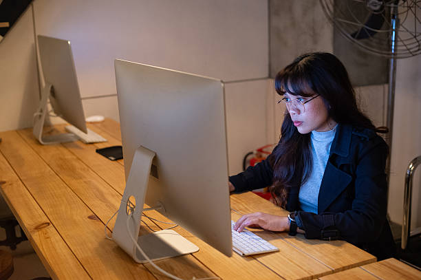 istock Young Asian woman diligently working on her computer with intense focus, typing on a keyboard at a wooden desk in a contemporary office setting. 2232717379