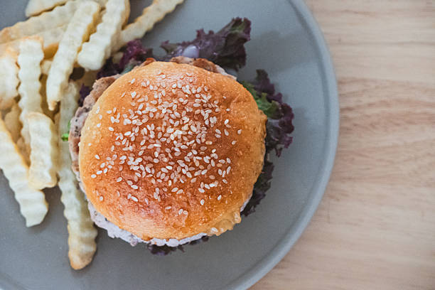 A close-up, top-down view of a delicious homemade burger topped with sesame seeds, served alongside crinkle-cut french fries on a grey plate, set on a light wooden table.