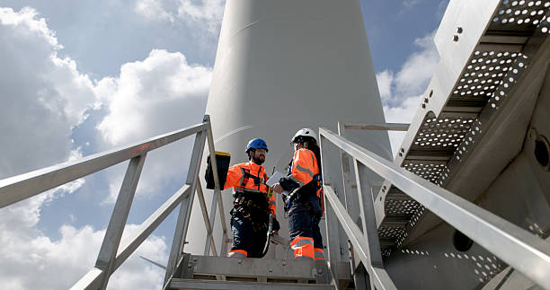 istock Two male and female technicians in safety gear, including helmets and safety harnes working for maintenance , Quality check under a large wind turbine 2232720607