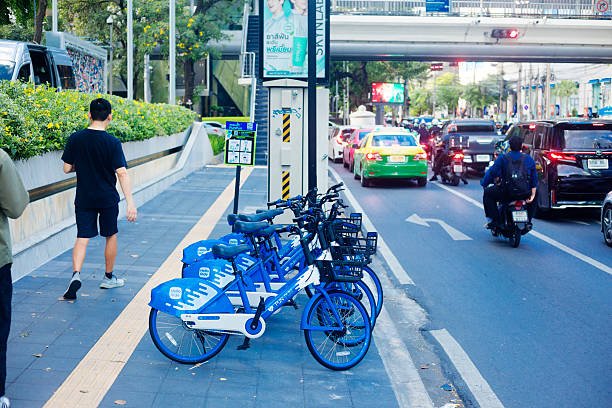 Parked blue city sharing bicycles on sidewalk in Bangok downtown at afternoon.  Scene is  at sidewalk of Wittayu Road near intersection with Phloen Chit Road. Behind bicycles is information sign how to unlock and rent  the bicycles. A man is walking along sidewalk. View from south to north direction. In background is intersection and on street are cars  and several motorcycle drivers. In background is a footbridge over the street