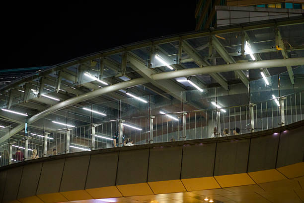 Walking people on modern footbridge at Terminal 21 in Bangkok downtown. Shopping mall Terminal 21 and BTS skytrain station and footbridge are connected at intersection of Thanon Asok and Sukhumvit Road. People are walking in both directions. View from parking driveway exit at side of mall.