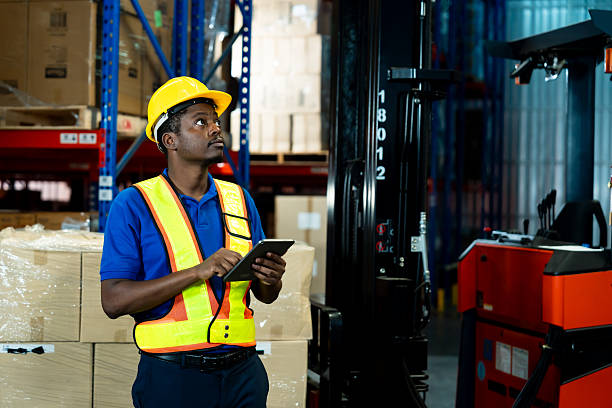 istock African male warehouse worker wearing helmet and safety vest using digital tablet while checking packages beside shelves and pallet rack in storage factory, inside logistics warehouse. 2232737414