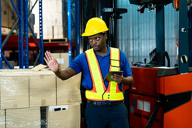 istock Adult African male warehouse worker explaining logistics tasks using digital tablet, standing in industrial storage area, wearing hard hat and vest, focusing on inventory coordination. 2232737419