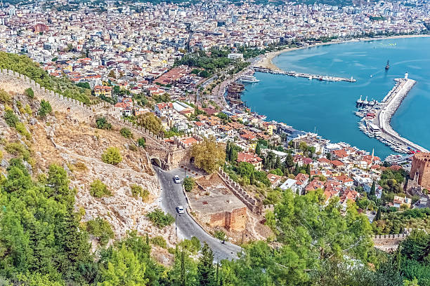A panoramic view of Alanya harbor from the Alanya Castle, showcasing the city below and the coastline stretching along the Mediterranean Sea. The Red Tower and historic fortifications are visible in the scene