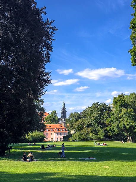 Beautiful park in Weimar, Germany featuring green grass, tree-lined pathways, and historic architecture in the background. People relax under the sunny sky creating a serene and welcoming atmosphere.
