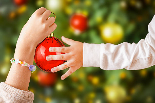 Close-up of two children's hands holding a shiny red Christmas ornament while decorating a festive Christmas tree. One child wears a colorful bead bracelet, adding a playful detail to the joyful holiday scene. Perfect image for Christmas, family traditions, and festive celebrations.