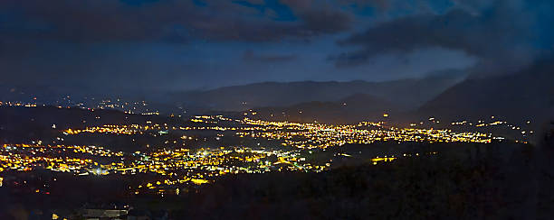 Wide-angle night panorama of a city glowing with golden lights, surrounded by dark hills and mountains under a cloudy sky. The illuminated streets and neighborhoods form a striking contrast against the moody evening atmosphere, highlighting urban life in harmony with nature.