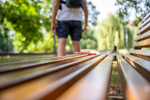 Seen from behind, a man in a white T-shirt and shorts walks away from a park bench. His smartphone rests on the bench, set against a tranquil park backdrop of green foliage and diffused sunlight.