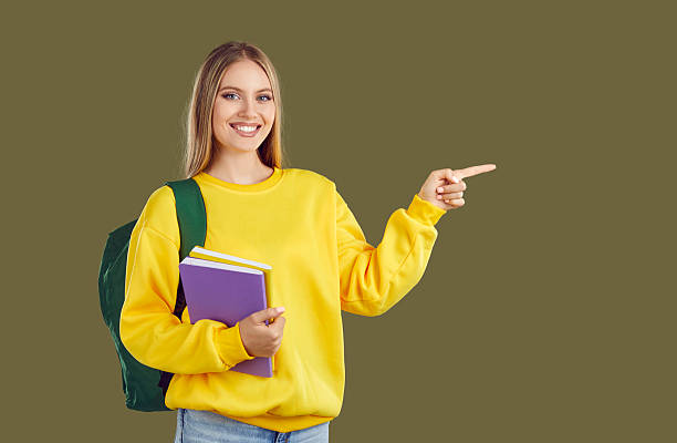 istock Happy student girl with backpack and copybooks pointing finger on copy space on khaki background. 2232764295