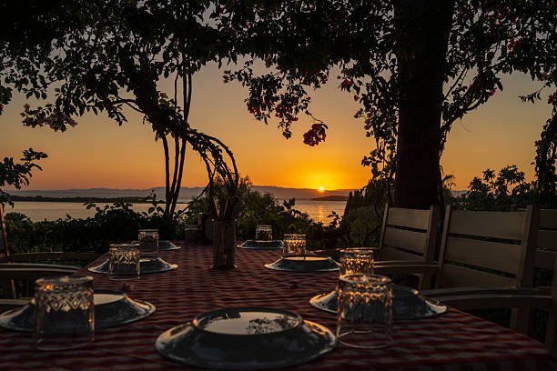 A beautifully set outdoor dinner table with checkered red cloth, glasses, and plates glowing under the warm sunset light, overlooking the tranquil sea. Perfectly capturing the essence of summer evenings by the coast.
