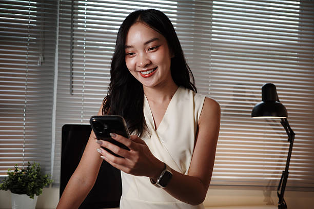 istock Young Asian businesswoman smiling while using her smartphone in a modern office, surrounded by technology and a professional atmosphere 2232781454