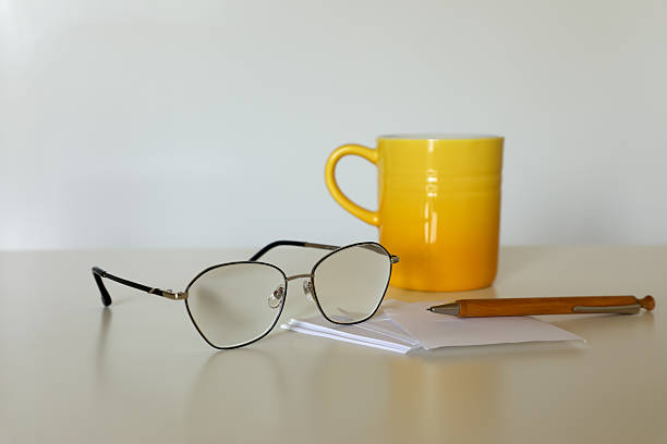 istock Eyeglasses, a wooden ballpoint pen, and a gradient yellow ceramic mug rest on folded white paper on a light-colored surface. The minimalist setup creates a cozy workspace atmosphere. 2232808196