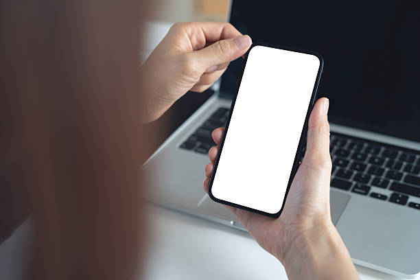 istock Blank white screen cell phone mockup. Close up, business woman using smartphone on office table during working on laptop computer, empty space for social media marketing or mobile app design, over shoulder view 2232809416