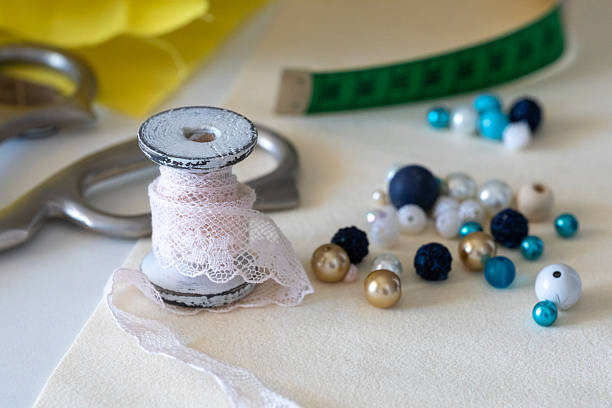 Crafting essentials close-up: pink lace, pearl, gold, blue beads, and sewing tools on cream. Ideal for handmade projects & creative hobbies.