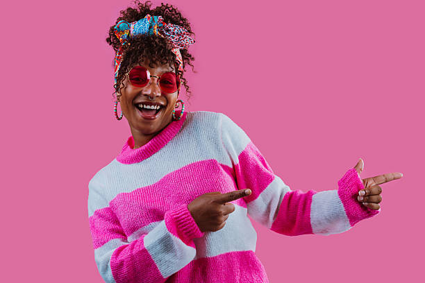 Fashion influencer wearing pink sunglasses and a cozy sweater, laughing joyfully while pointing sideways with index fingers against a vibrant pink background, radiating cheerful energy