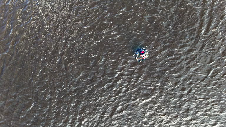 From a steady overhead drone, a lone diver swims beside a bright marker buoy on textured open water, suggesting training, alert awareness, and measured endurance in a minimalist scene.