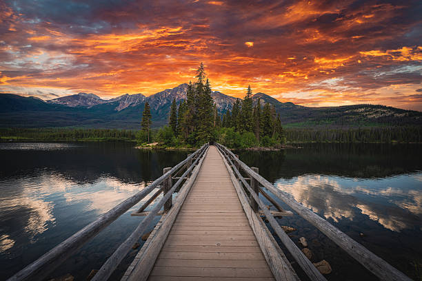 Dramatic sunset over Pyramid Lake with the wooden bridge leading to Pyramid Island in Jasper National Park, Alberta, Canada. The fiery sky reflects in the calm waters, framed by evergreen forests and rugged peaks of the Canadian Rockies. A breathtaking and iconic Canada travel destination perfect for themes of nature, adventure, exploration, and scenic landscapes.