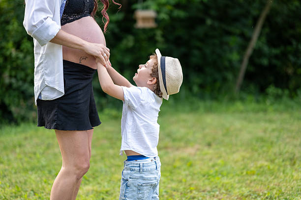 An american shot photo of a young child tenderly touches a pregnant woman's belly in a serene outdoor setting, symbolizing familial love and anticipation. The woman, in a stylish black skirt and white shirt, and the child, in a white t-shirt and denim shorts, exude warmth and connection amidst lush greenery.