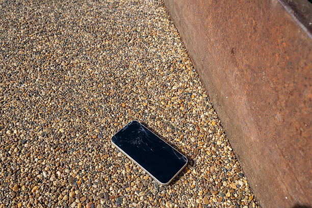 Modern mobile phone with shattered screen abandoned on a gravel surface next to a rusty metal sheet, illustrating concepts of damage, loss, and obsolescence
