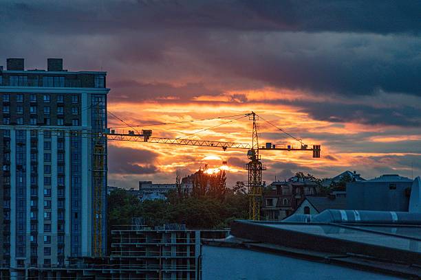 Urban scene with construction cranes, buildings under construction, scaffolding, unfinished facades, sunset, clouds, sky, long shadows, power lines, elevated roadway, architectural details a