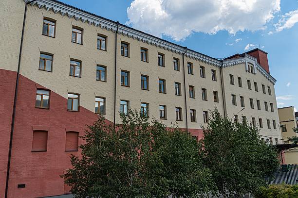 istock Multi-story, red-hued building with vertical windows and shutters, contrasting lighter sections, architectural detail or gap, lush green trees, daytime, ample natural light, scattered clouds, blue sk 2232864497