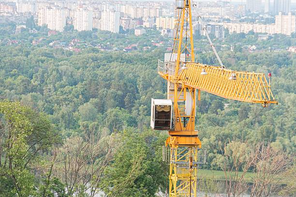 istock Yellow construction crane on an elevated area, possibly a hillside, with residential structures below The crane has multiple sections, a cab, and a long jib Soft lighting conditions suggest early m 2232865393
