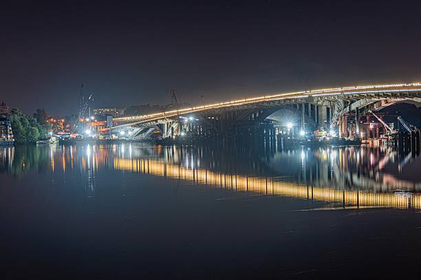 istock Night scene at a harbor featuring a glowing bridge with multiple arches spanning still water, two moored boats, warm yellow reflection, soft lighting, calm atmosphere, minimal activity, dreamy qualit 2232867029