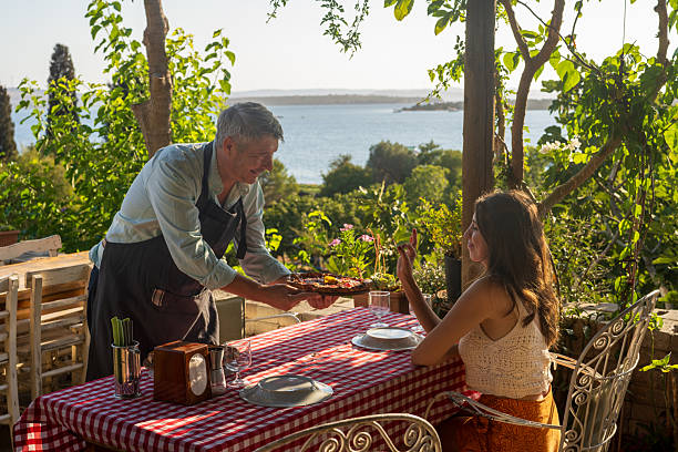 In a charming countryside setting with a breathtaking sea view, a small business owner proudly serves his freshly baked, wood-fired pizza to a guest. The warm light, rustic décor, and authentic gesture highlight the soul of local gastronomy and the passion behind small family-run restaurants.