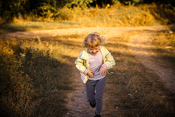 A young child in a light jacket dashes along a dirt trail surrounded by golden grass and green foliage. The scene captures the carefree spirit of childhood in a serene outdoor setting.
