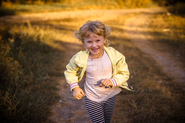 A young child in a light jacket dashes along a dirt trail surrounded by golden grass and green foliage. The scene captures the carefree spirit of childhood in a serene outdoor setting.