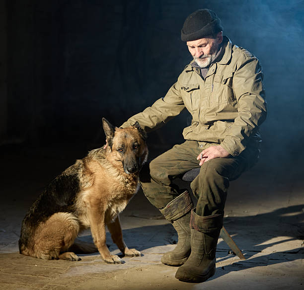 Senior man with grey beard dressed in shabby old clothes and in knitted hat is spending time with his dog female german shepherd. Shooting in abandoned factory