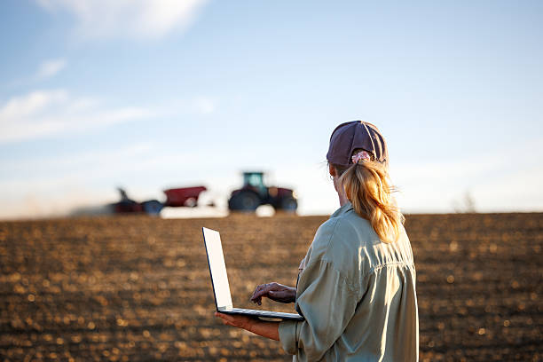 istock Woman farmer with laptop analyzing agricultural data in front of tractor with seed drill sowing crops at field 2232890004