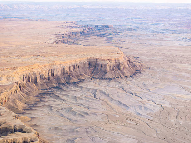 Aerial view of an isolated flat-topped mesa rising from Utah’s vast desert badlands, with dramatic cliffs, layered rock formations, and expansive barren terrain stretching into the distance.