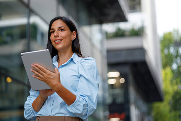 istock Young businesswoman smiling while looking away, using a digital tablet in an urban environment filled with modern architecture 2232902387